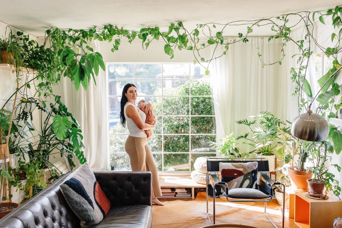 Ashlee standing in a sunlit, plant-filled living room cradling a swaddled newborn — an editorial portrait of warm, rooted presence