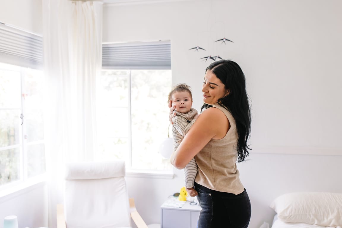 Ashlee McKenzie standing by a window holding a baby close, soft daylight filling the nursery